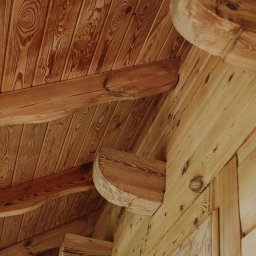 Łukasz Drężewski - Drewniany sufit i konstrukcja z belek, widoczne słoje drewna. Ciepłe oświetlenie podkreśla naturalny charakter materiału. Detal architektoniczny z bliska.
