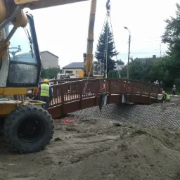 Żuraw ustawia przęsło drewnianego mostu dla pieszych nad przygotowanym terenem z umocnionym brzegiem, widoczni pracownicy w kamizelkach odblaskowych.