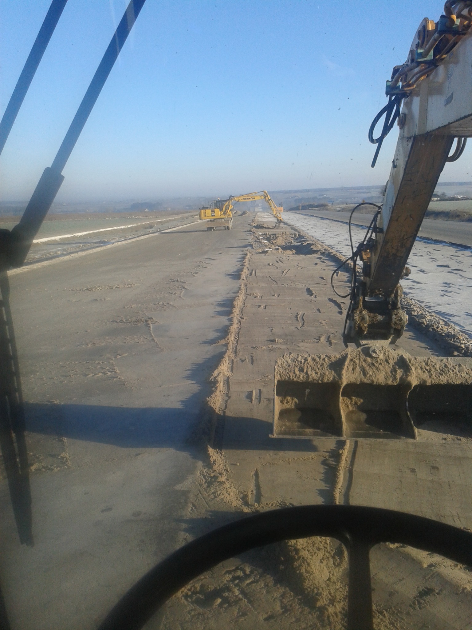 Widok z kabiny koparki na utwardzany teren, w oddali druga koparka, błękitne niebo w tle.