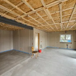 Surowy stan wnętrza w trakcie remontu: widoczne ocieplenie wełną mineralną pod sufitem z drewnianą konstrukcją, ściany wykończone płytami gipsowo-kartonowymi, betonowa posadzka oraz krzesło...