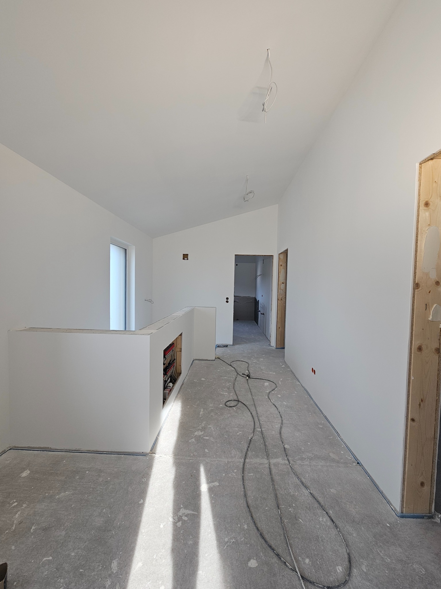 Wnętrze w trakcie prac wykończeniowych: świeżo pomalowane na biało ściany, surowa drewniana ościeżnica, przewody elektryczne na podłodze, widok na dalsze pomieszczenia.