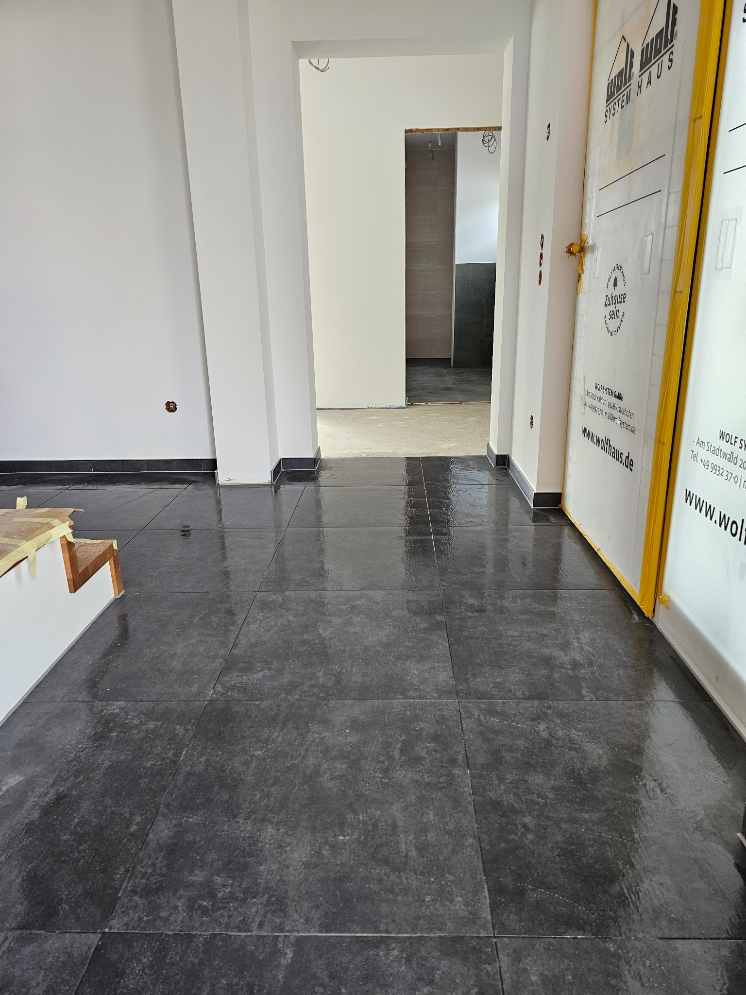 Wypolerowana, ciemna podłoga z płytek w nowym budynku, z widocznymi fragmentami schodów zabezpieczonych kartonem, białe ściany z czarną listwą przypodłogową i zabezpieczone drzwi z logo firmy...