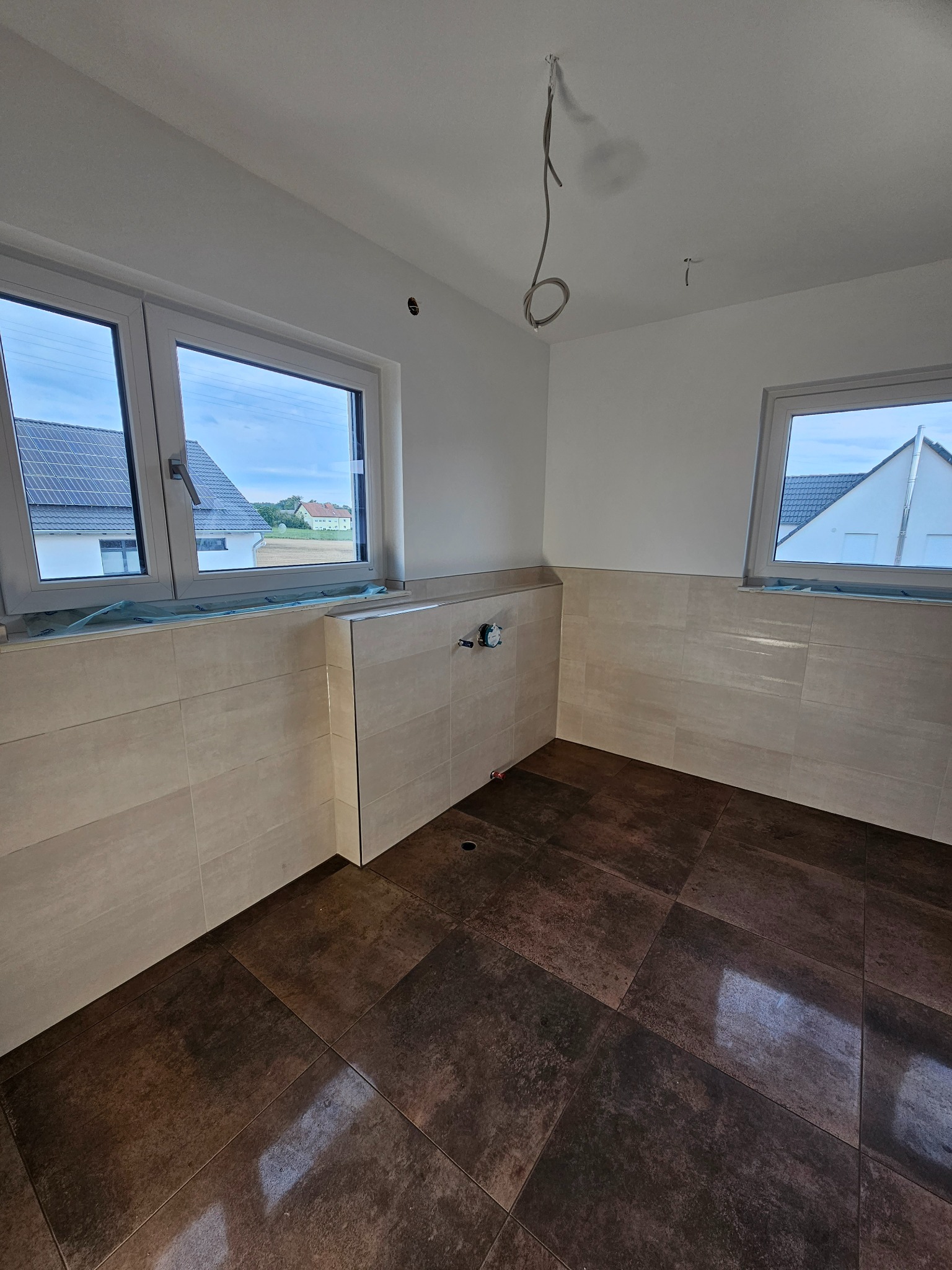 Wnętrze łazienki w trakcie prac wykończeniowych: ułożone płytki ścienne w beżowym odcieniu i podłogowe w ciemnym brązie, widoczne wyprowadzenia instalacji wodnej, okna zabezpieczone folią ochronną,...