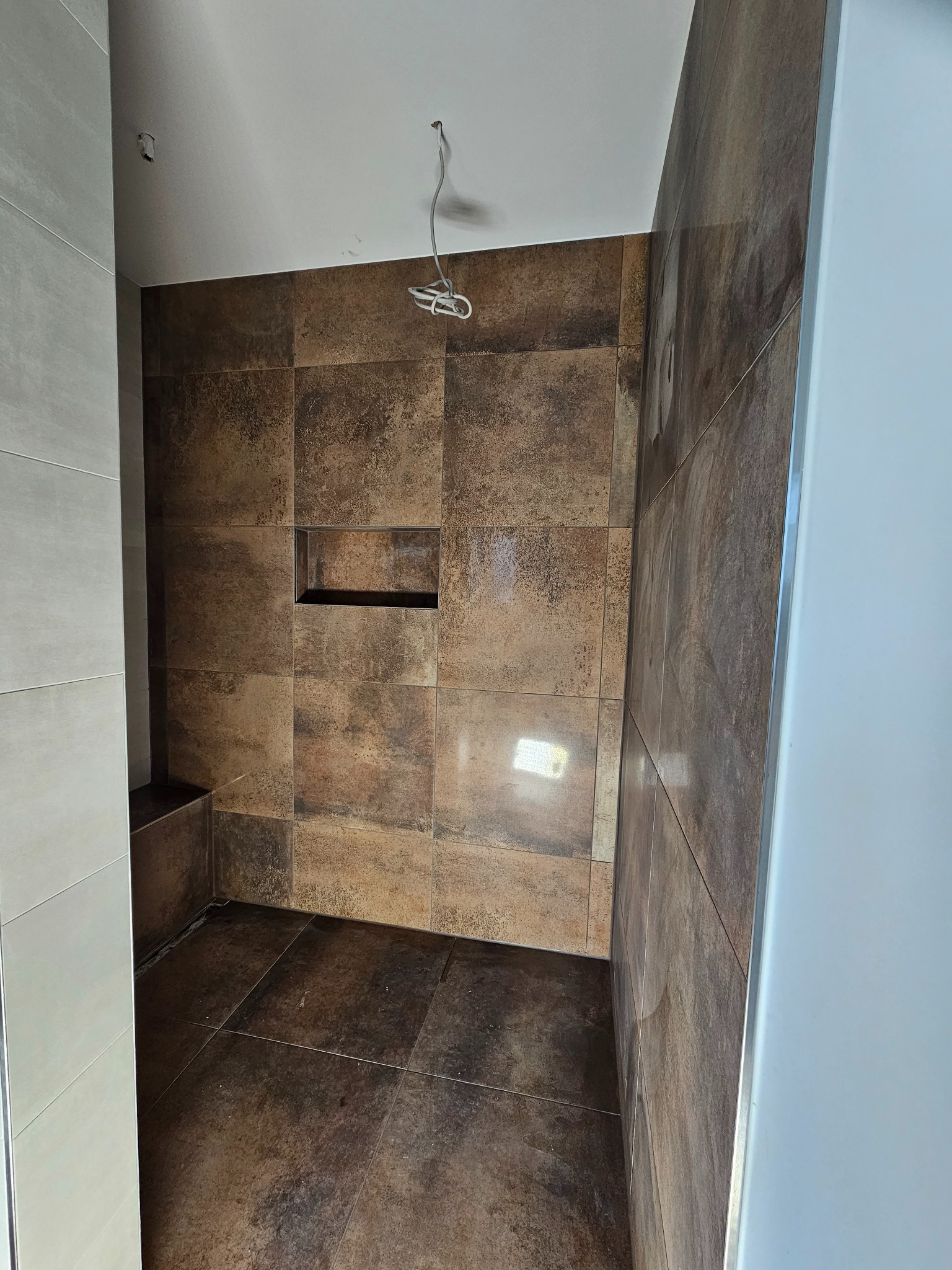 Wnętrze kabiny prysznicowej w trakcie wykańczania, widoczne brązowe płytki imitujące kamień na ścianach i podłodze, w ścianie wnęka, wystające przewody elektryczne, brak armatury.