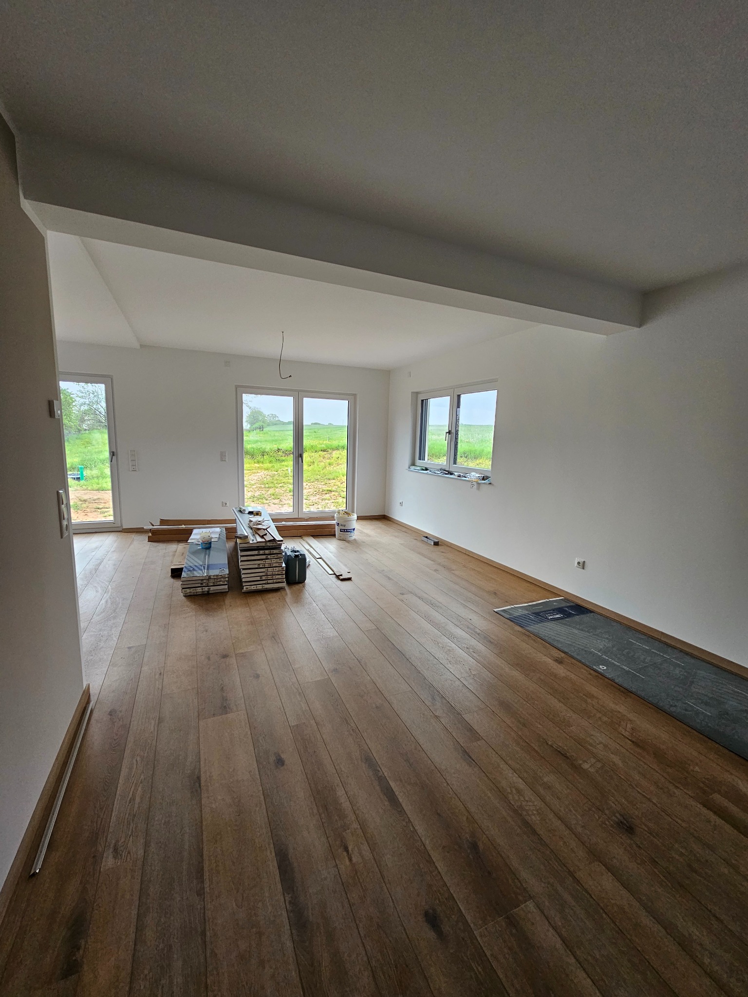 Wnętrze pokoju z drewnianą podłogą w trakcie wykańczania, widoczne okna z widokiem na zewnątrz, białe ściany, przygotowane panele podłogowe i wiaderko z farbą.