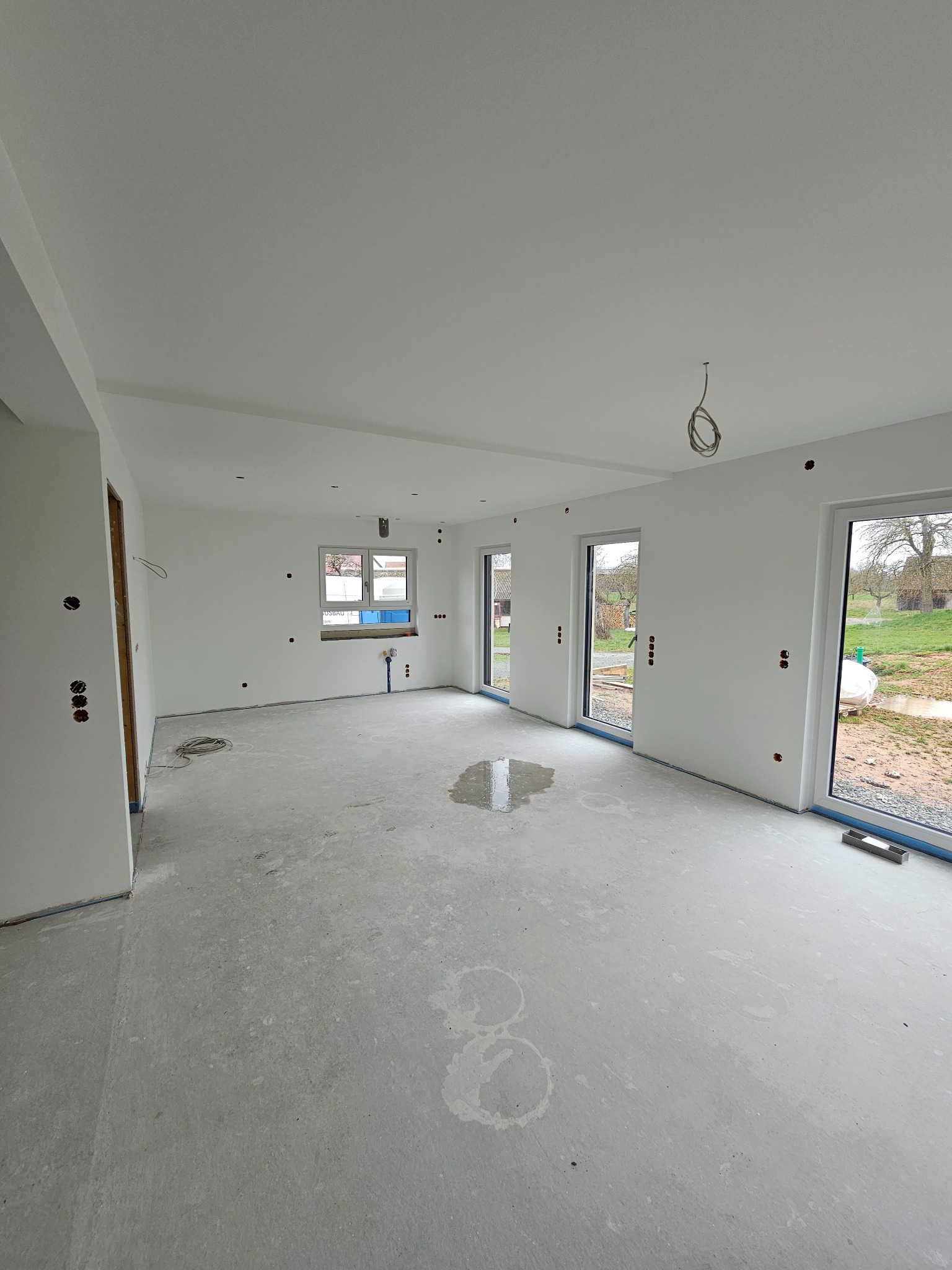 Wnętrze nowego domu w stanie deweloperskim, z białymi ścianami, surową betonową podłogą z widocznymi plamami wody, przygotowane do dalszych prac wykończeniowych, widoczne puszki elektryczne...
