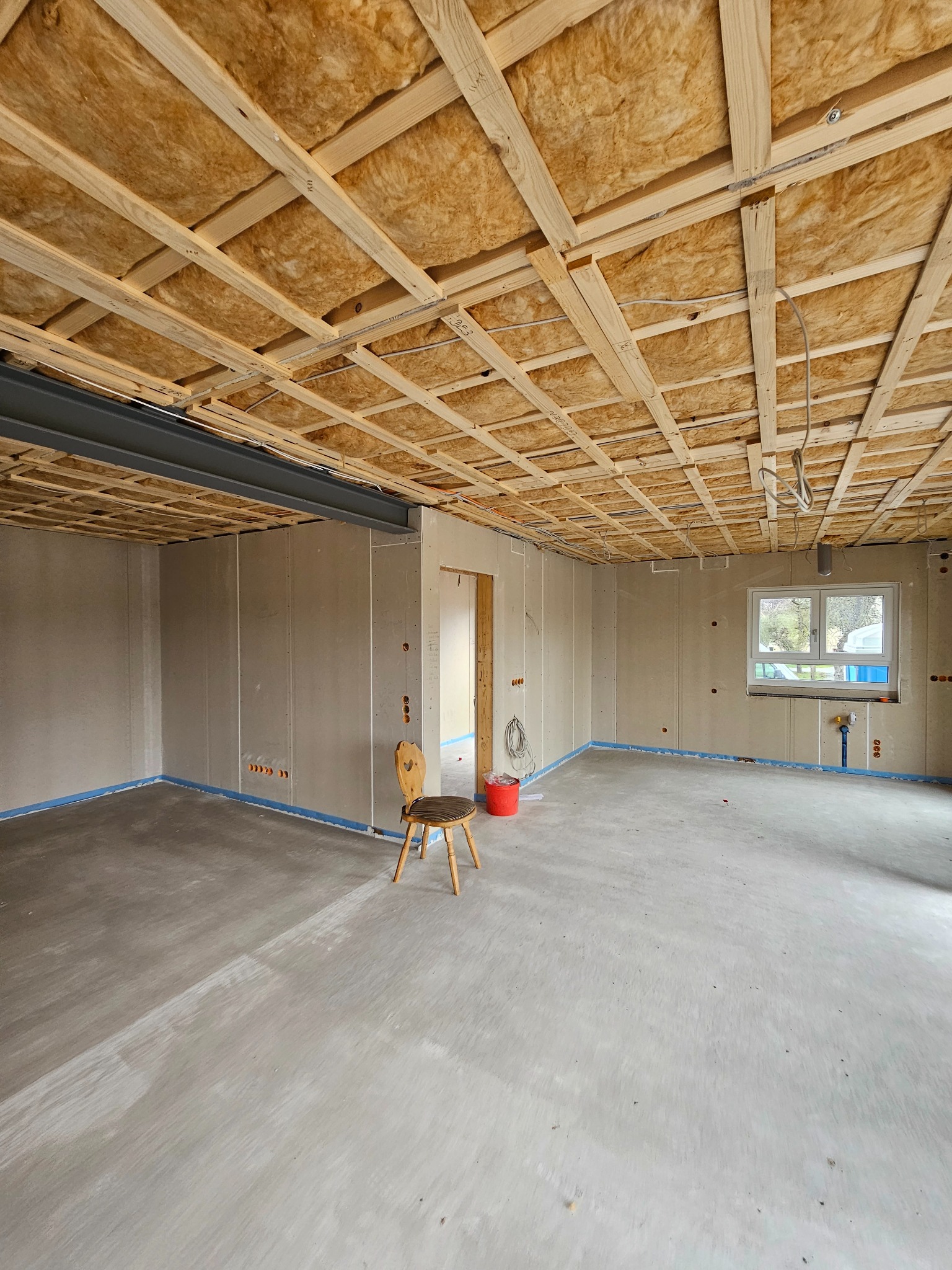 Surowy stan wnętrza w trakcie remontu: widoczne ocieplenie wełną mineralną pod sufitem z drewnianą konstrukcją, ściany wykończone płytami gipsowo-kartonowymi, betonowa posadzka oraz krzesło...