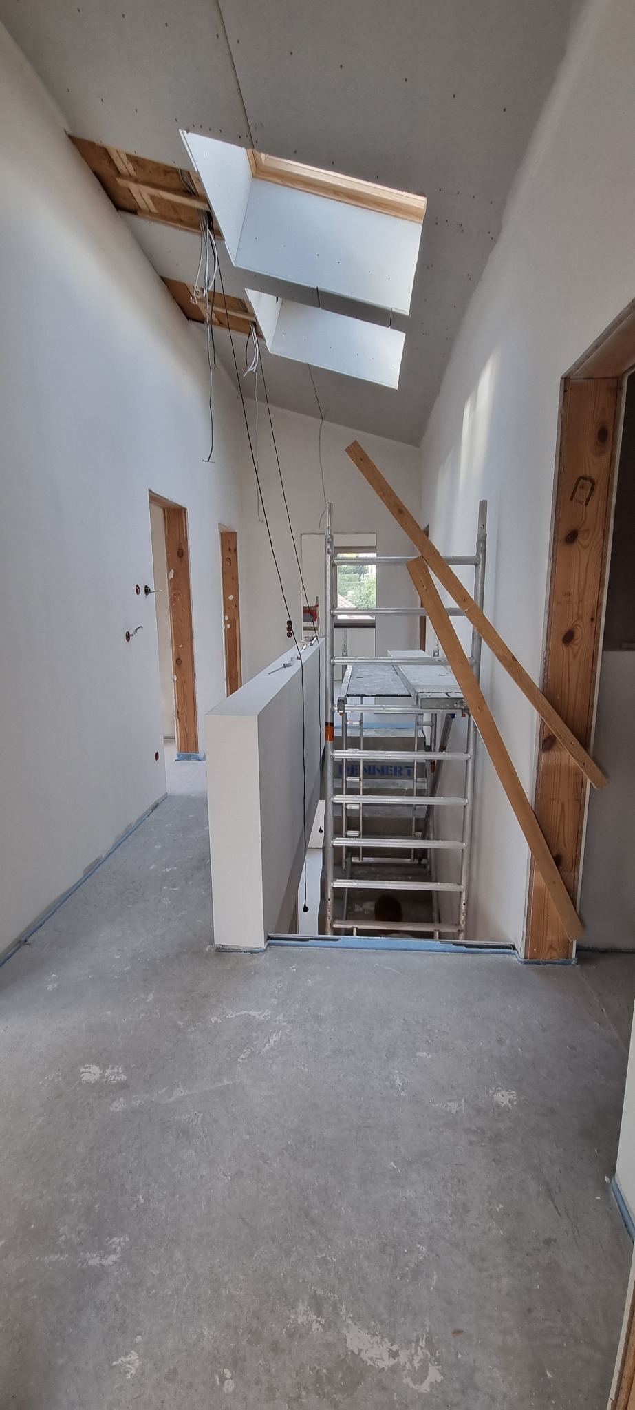Wnętrze w trakcie remontu: surowe ściany, widoczne okna dachowe z wystającymi przewodami, metalowe rusztowanie z drewnianymi elementami opartymi o framugę drzwi, surowy beton na podłodze.