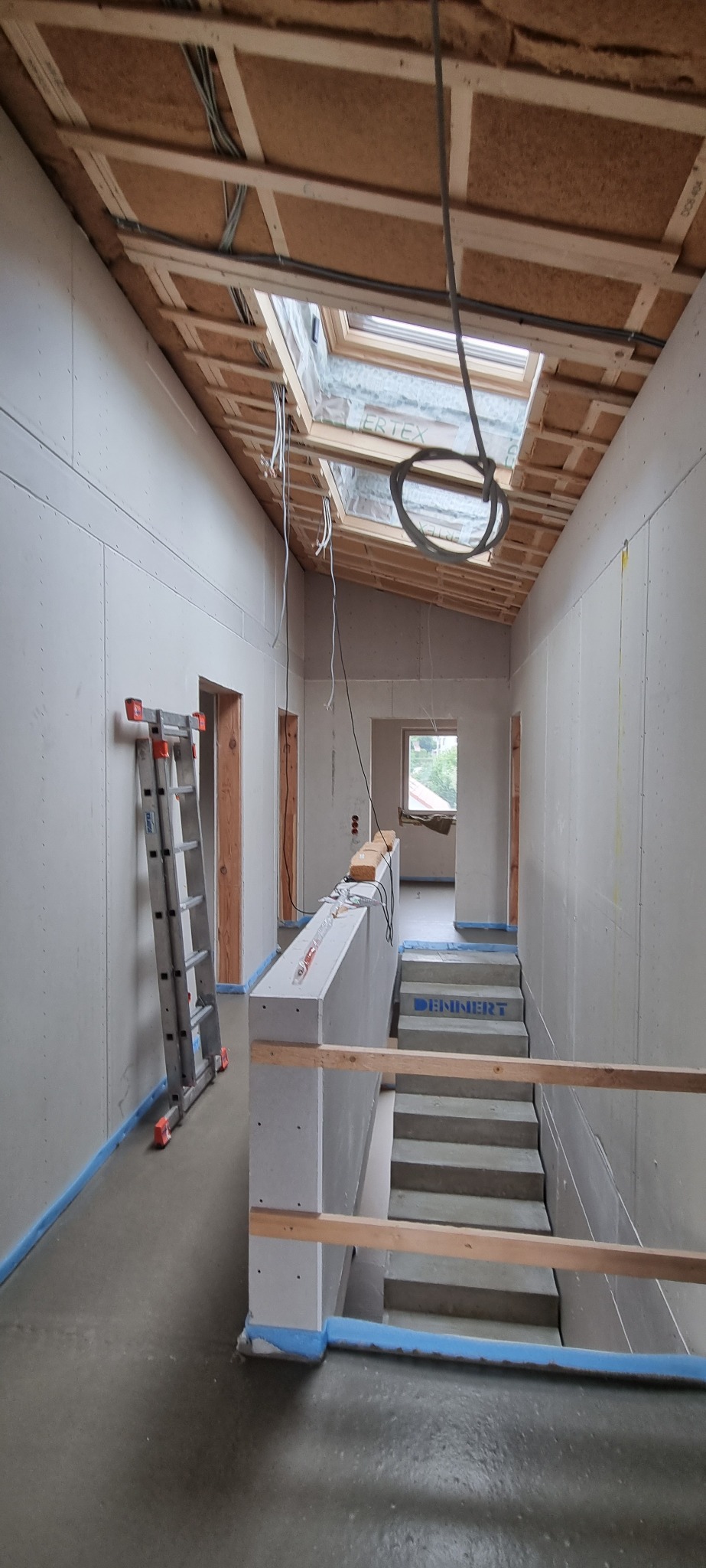 Korytarz w trakcie wykańczania: ściany pokryte płytami gipsowo-kartonowymi, widoczne kable zwisające z sufitu z oknem dachowym, schody betonowe z zabezpieczeniem folią, drabina oparta o ścianę...