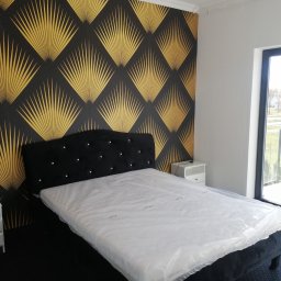 Krzysztof Janas firma ogólnobudowlana - Sypialnia z łóżkiem z czarnym, aksamitnym zagłówkiem z kryształkami na tle ściany z czarno-złotą tapetą w geometryczne wzory, lampa wisząca i biała szafka nocna.