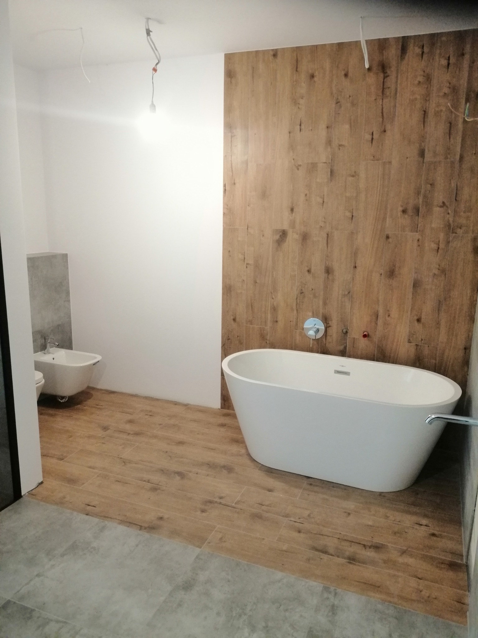 Nowoczesna łazienka w trakcie wykańczania: wolnostojąca wanna, bidet i płytki imitujące drewno na podłodze i ścianie, widoczne niedokończone instalacje elektryczne.
