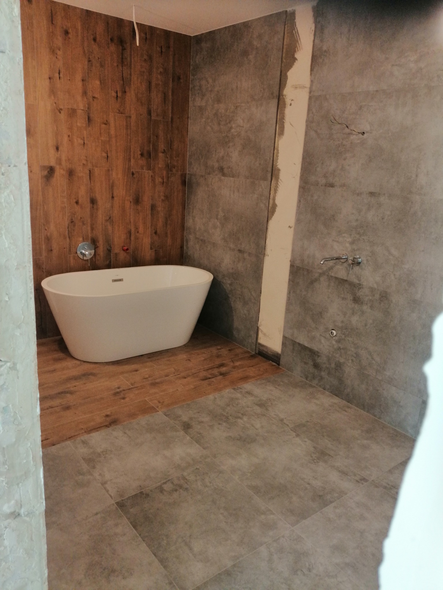 Nowoczesna łazienka w trakcie remontu: biała wanna wolnostojąca na tle drewnopodobnych paneli, szare płytki na podłodze i ścianach, widoczne ślady prac wykończeniowych.