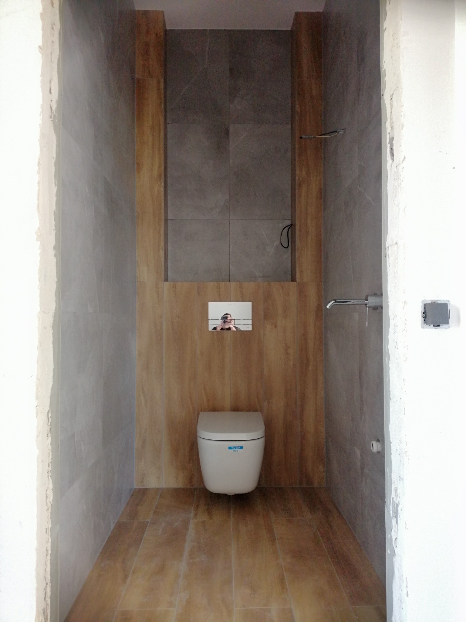 Wnętrze łazienki w trakcie remontu, widoczna podwieszana toaleta, fragment lustra z odbiciem osoby, szare i drewnopodobne płytki na ścianach i podłodze, surowy stan wykończenia przy framudze drzwi.