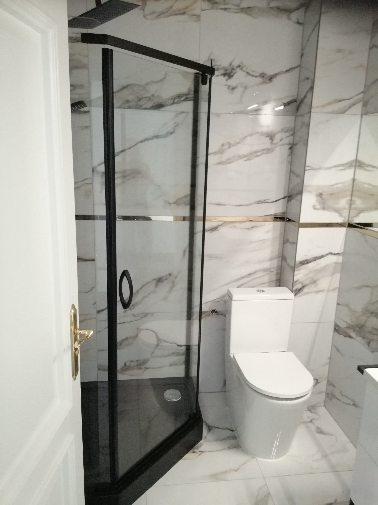 Nowoczesna łazienka z kabiną prysznicową z czarnymi ramami, białą toaletą i marmurowymi płytkami na ścianach i podłodze.
