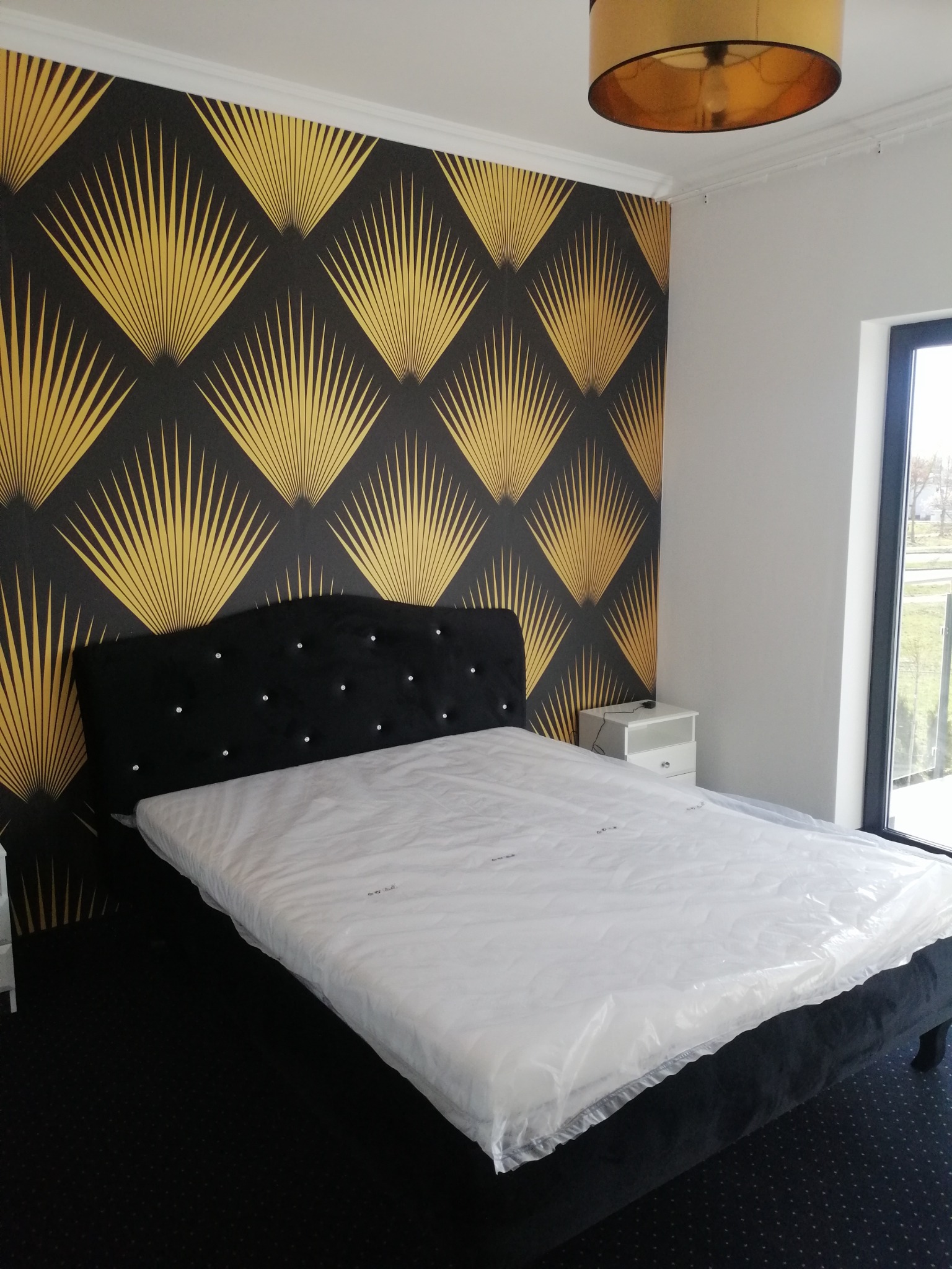 Sypialnia z łóżkiem z czarnym, aksamitnym zagłówkiem z kryształkami na tle ściany z czarno-złotą tapetą w geometryczne wzory, lampa wisząca i biała szafka nocna.