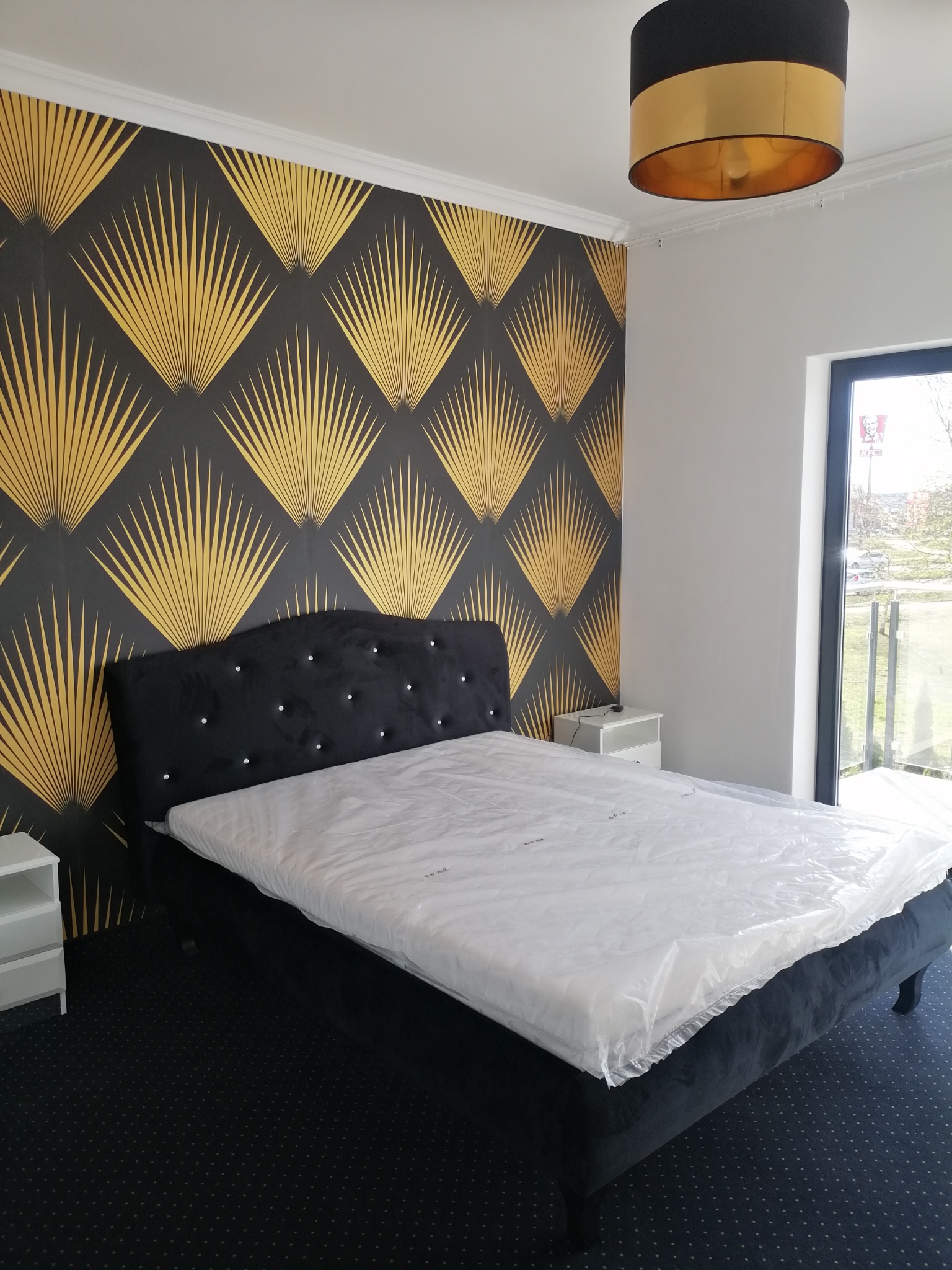 Sypialnia z czarnym łóżkiem z kryształkami na wezgłowiu, złotą tapetą w geometryczne wzory i czarnym dywanem w kropki, widok na zewnątrz przez okno.