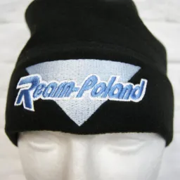 Czarna czapka z polaru z haftowanym napisem 'Team-Poland' w kolorze niebieskim, umieszczona na białej, styropianowej głowie manekina na tle białej ściany z cegieł.