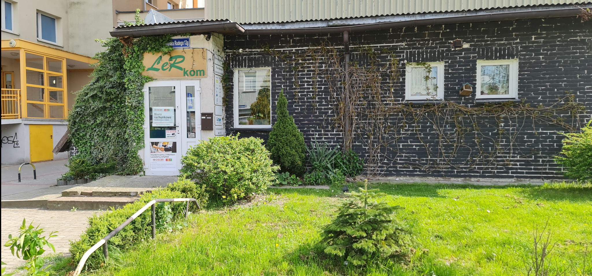 Wejście do biura rachunkowego z nazwą 'LeRkom' porośnięte bluszczem, czarna ceglana ściana, okna z firankami i zadbany trawnik z krzewami.