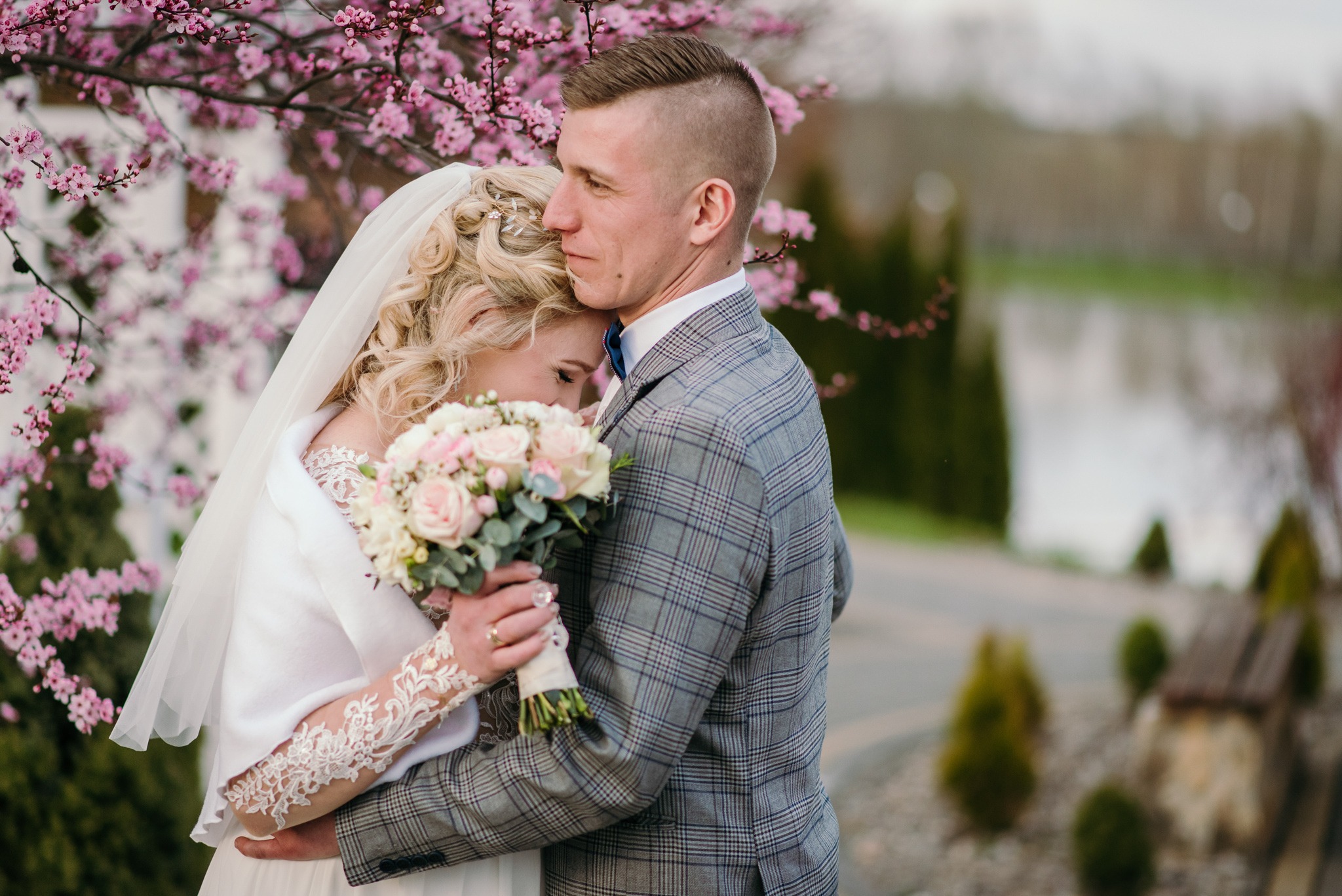 Para młoda w objęciach pod kwitnącym drzewem wiśni, panna młoda z bukietem różowych i kremowych kwiatów, on w szarej kraciastej marynarce i muszce.