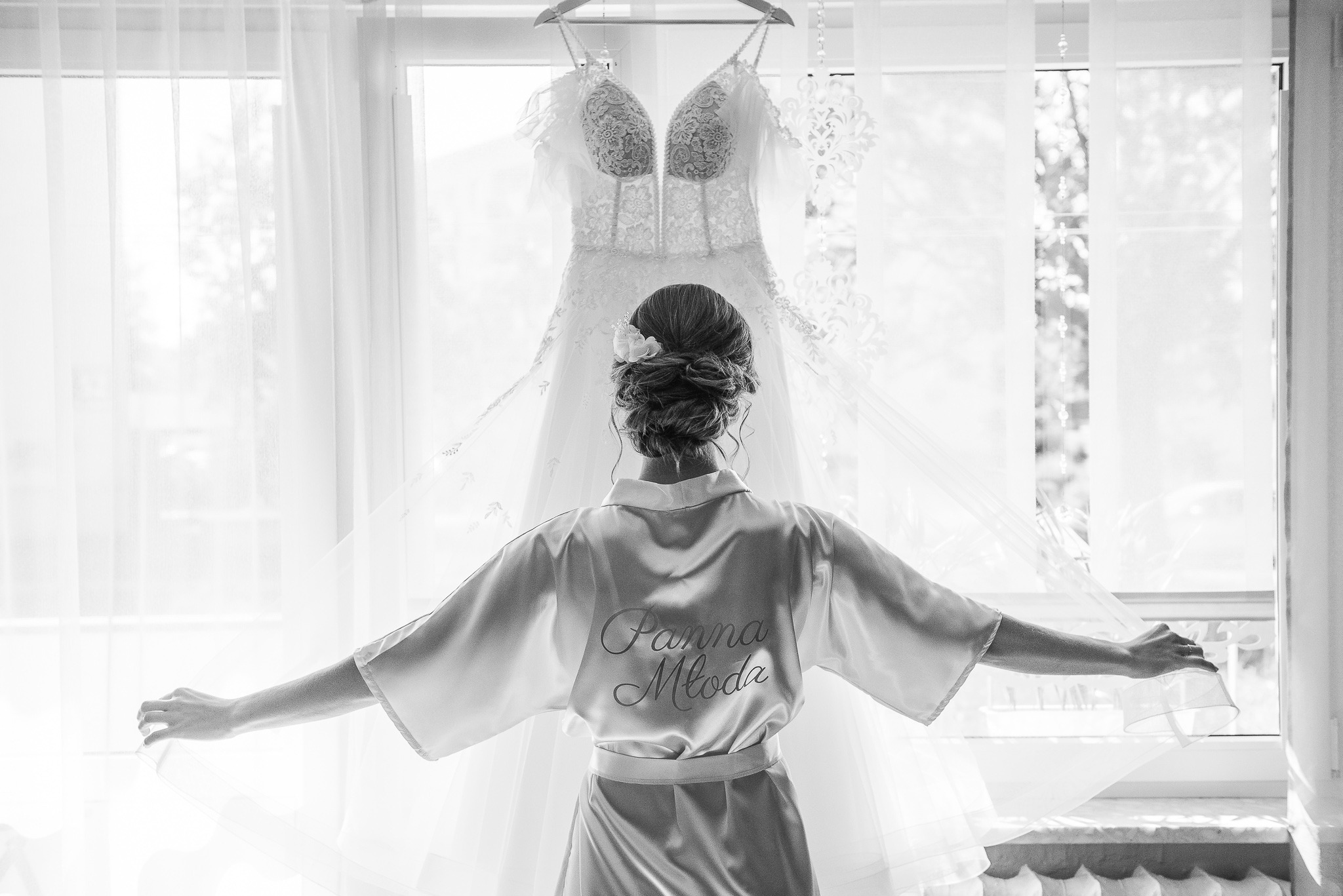Panna młoda w szlafroku z napisem 'Panna Młoda' stoi tyłem, rozkładając ręce na tle okna i wiszącej sukni ślubnej. Scena w czerni i bieli.