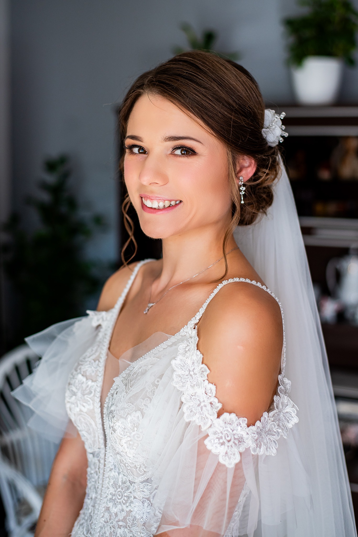 Uśmiechnięta panna młoda w białej sukni z koronką, z delikatnym welonem i kwiatową ozdobą we włosach, pozuje w jasnym wnętrzu z rośliną w tle.