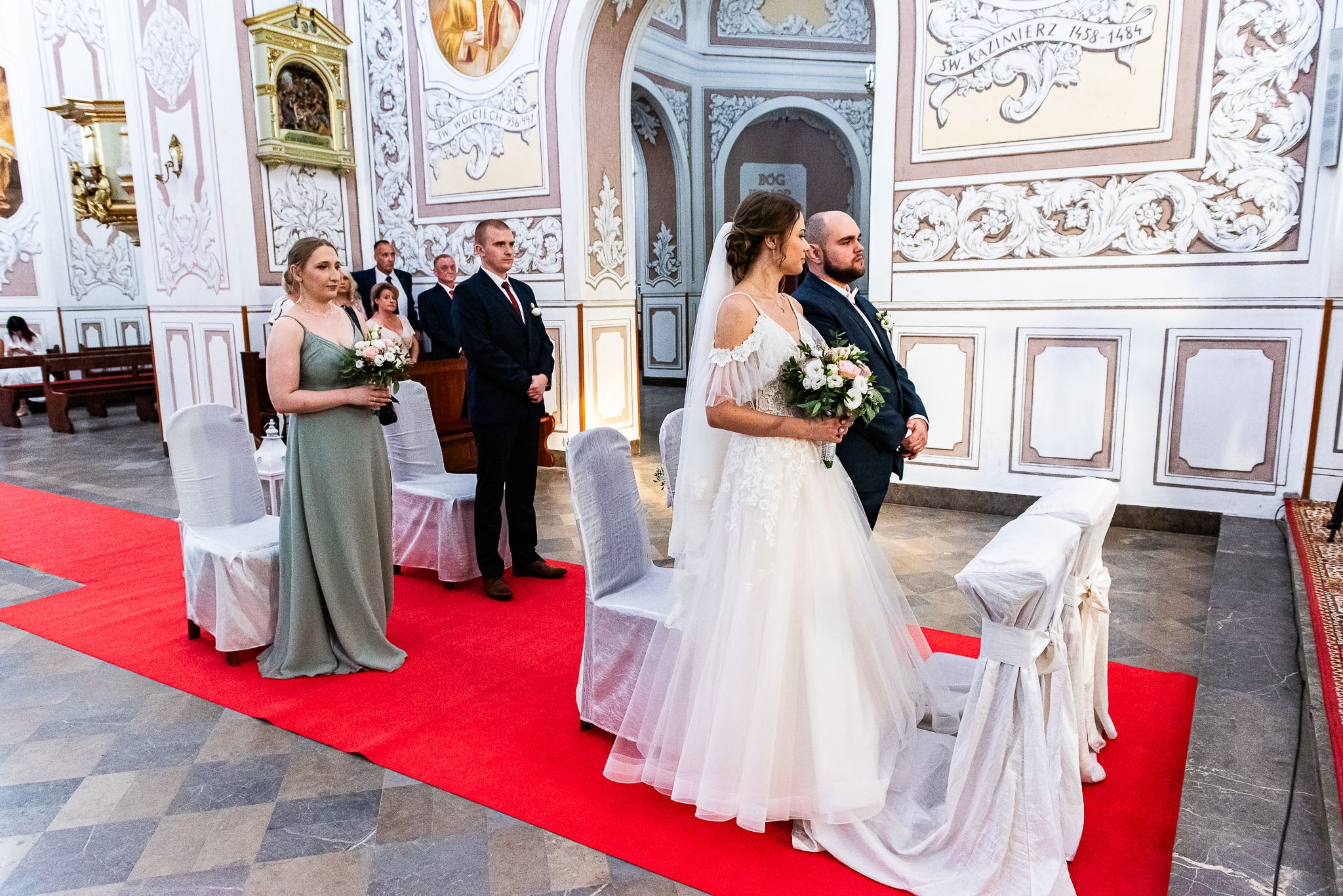 Para młoda w kościele, panna młoda w białej sukni z bukietem, obok drużba i goście w tle, czerwony dywan, bogato zdobione ściany.