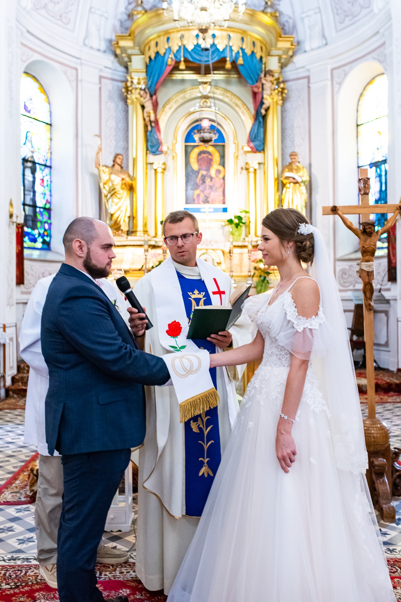 Uroczystość ślubna w kościele: panna młoda w białej sukni, pan młody w granatowym garniturze, ksiądz z mikrofonem i stułą z motywem obrączek.