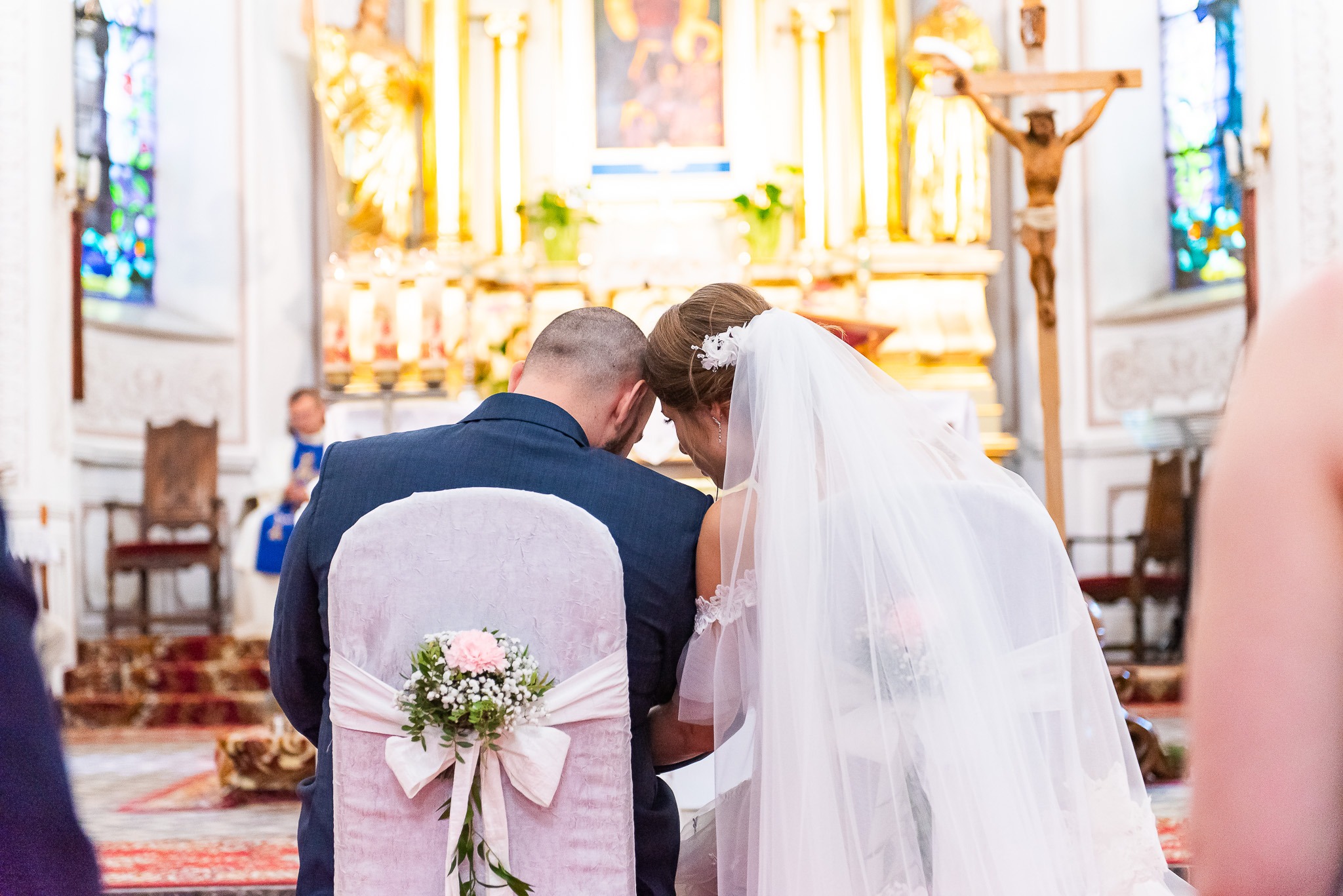 Para młoda w kościele podczas ceremonii ślubnej, siedząca na udekorowanych krzesłach z kwiatami, skierowana przodem do ołtarza. W tle widoczny ksiądz.