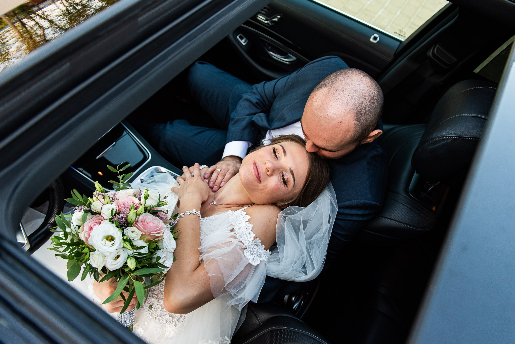 Para młoda w samochodzie z otwartym dachem: panna młoda w sukni z bukietem, pan młody w garniturze, romantyczny pocałunek. Ujęcie z góry.