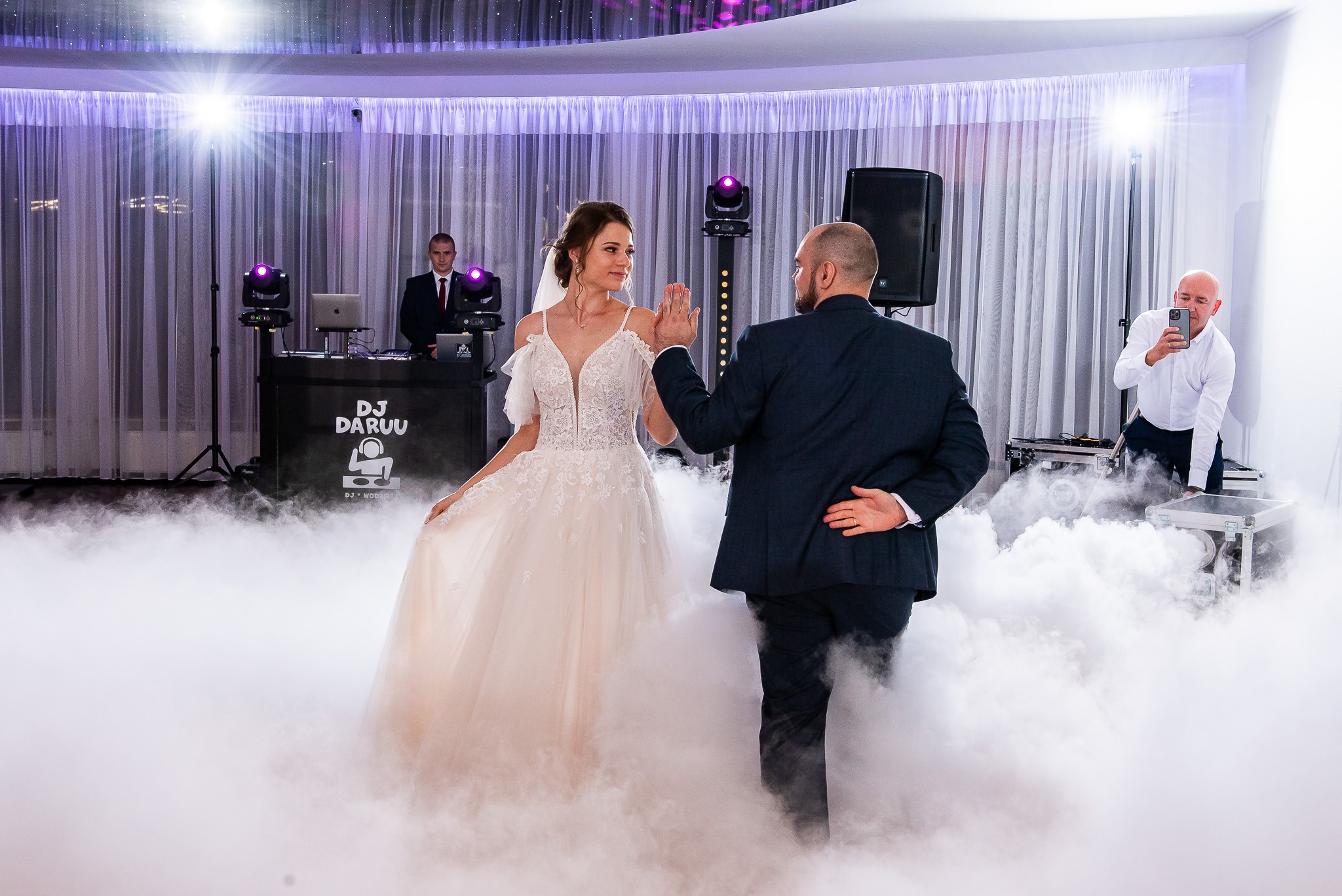 Para młoda w tańcu, otoczona dymem na parkiecie podczas wesela. W tle DJ i osoba filmująca telefonem. Dekoracje świetlne i białe zasłony.