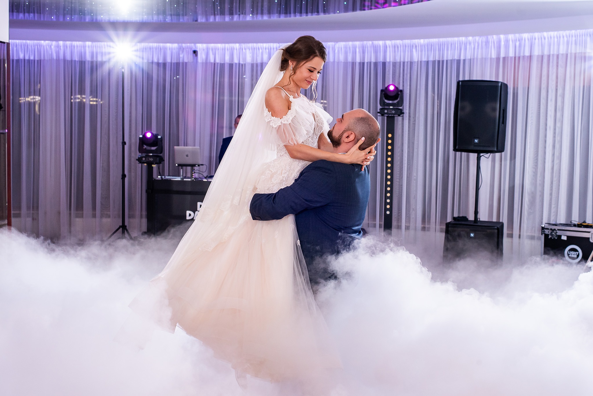 Uśmiechnięta panna młoda w objęciach pana młodego podczas pierwszego tańca w dymie, na tle sceny z oświetleniem i sprzętem DJ-skim.