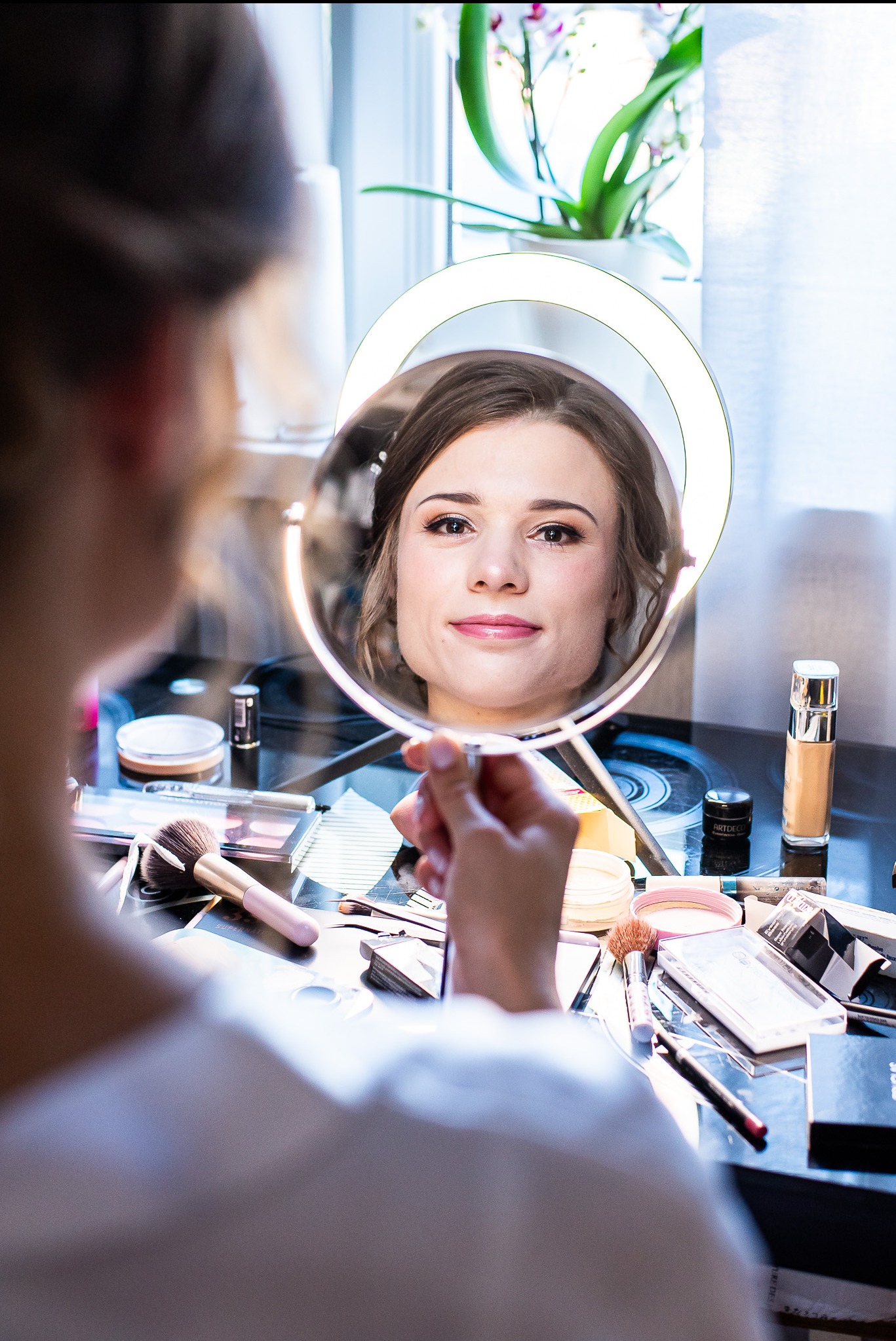 Uśmiechnięta kobieta w okrągłym lustrze z podświetleniem, otoczona kosmetykami na czarnym blacie, widziana zza ramienia drugiej osoby.