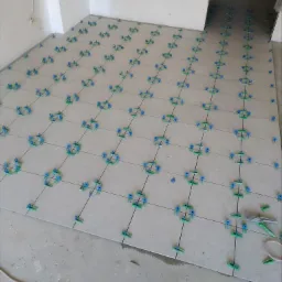 Układanie jasnych płytek podłogowych w trakcie prac, z widocznymi klipsami poziomującymi i krzyżykami dystansowymi, oraz narzędziem do cięcia płytek w prawym dolnym rogu.