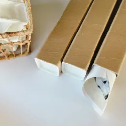 Trzy tuby z tektury zawierające wydruki, z widocznym fragmentem portretu psa na jednym z nich, obok plecionego koszyka z białymi wkładami, na jasnym tle.