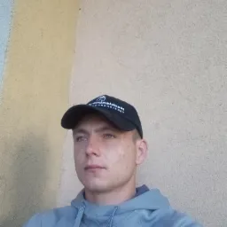 Portret młodego mężczyzny w czapce z daszkiem i szarej bluzie na tle ściany z nierówną, jasną fakturą.