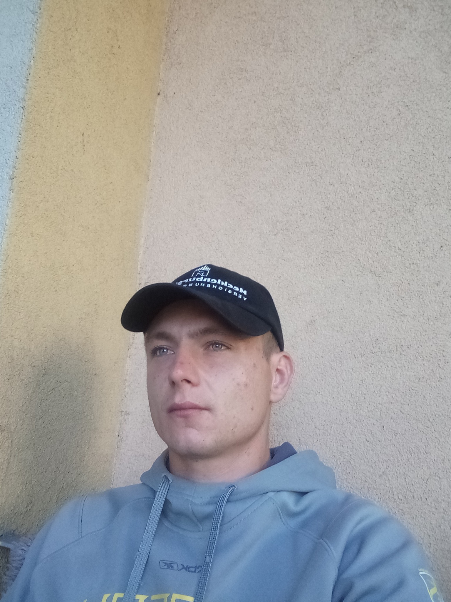 Portret młodego mężczyzny w czapce z daszkiem i szarej bluzie na tle ściany z nierówną, jasną fakturą.