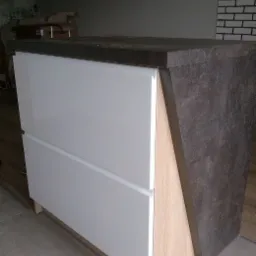 Nowoczesna szafka z białymi, lakierowanymi frontami szuflad i blatem imitującym ciemny kamień, z widoczną strukturą drewna na bocznych panelach, ustawiona na tle ściany z cegieł.