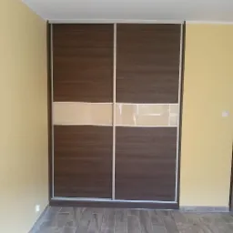 Dwudrzwiowa szafa przesuwna z brązowymi panelami imitującymi drewno i poziomym pasem z mlecznego szkła, w pomieszczeniu z ż&oacute;łtymi ścianami i szarą podłogą z paneli.