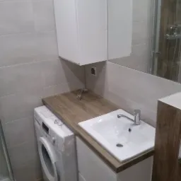 Mała łazienka z pralką obok szafki umywalkowej z drewnianym blatem, białe szafki wiszące i lustro.