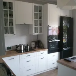 Jasna kuchnia z białymi szafkami z ciemnymi uchwytami, drewnianym blatem, czarną lodówką i telewizorem nad piekarnikiem.