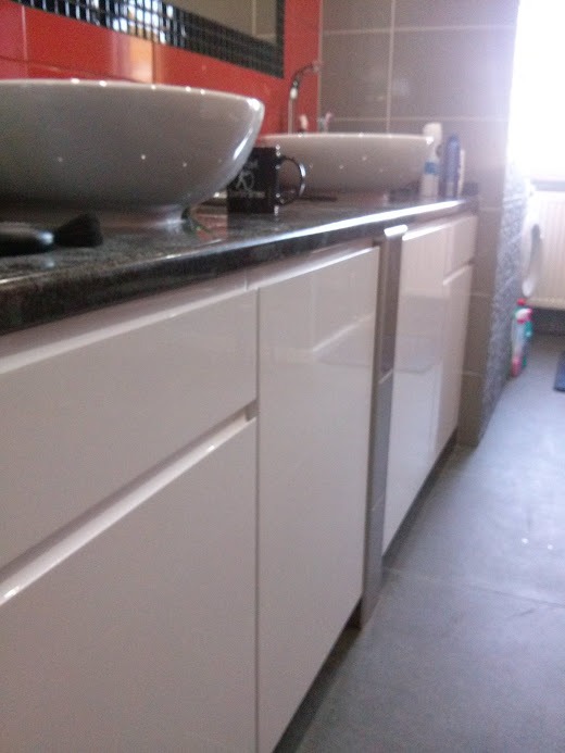 Łazienka z białymi szafkami na wymiar pod umywalkami, czarny blat i czerwona ściana z czarną mozaiką w tle.