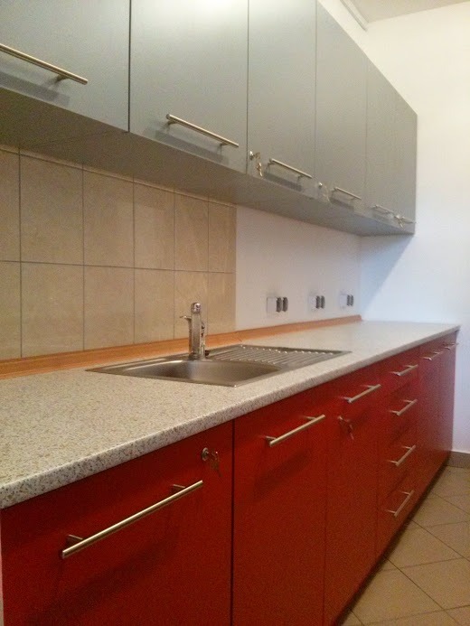 Nowoczesna kuchnia z szarymi szafkami wiszącymi i czerwonymi szafkami dolnymi, blatem w jasnym odcieniu i metalowym zlewozmywakiem. Widoczne długie, poziome uchwyty. Na ścianie beżowe płytki...