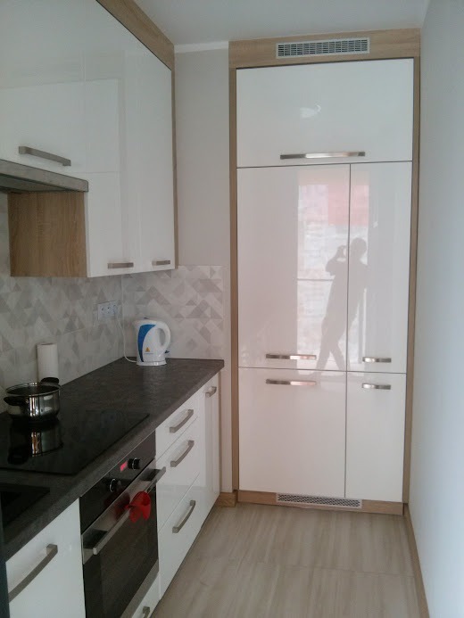 Nowoczesna, biała zabudowa kuchenna z błyszczącymi frontami, szarym blatem i akcentami w kolorze drewna, widoczny fragment płyty grzewczej z garnkiem, czajnik elektryczny i geometryczna mozaika...