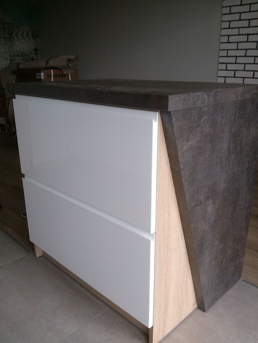 Nowoczesna szafka z białymi, lakierowanymi frontami szuflad i blatem imitującym ciemny kamień, z widoczną strukturą drewna na bocznych panelach, ustawiona na tle ściany z cegieł.