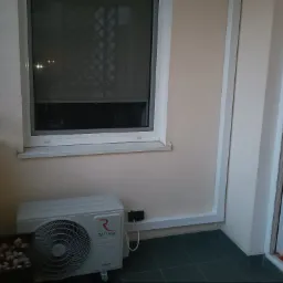 Jednostka zewnętrzna klimatyzatora Rotenso Ukura zamontowana na balkonie, widoczny kabel zasilający i doniczka z kamieniami.