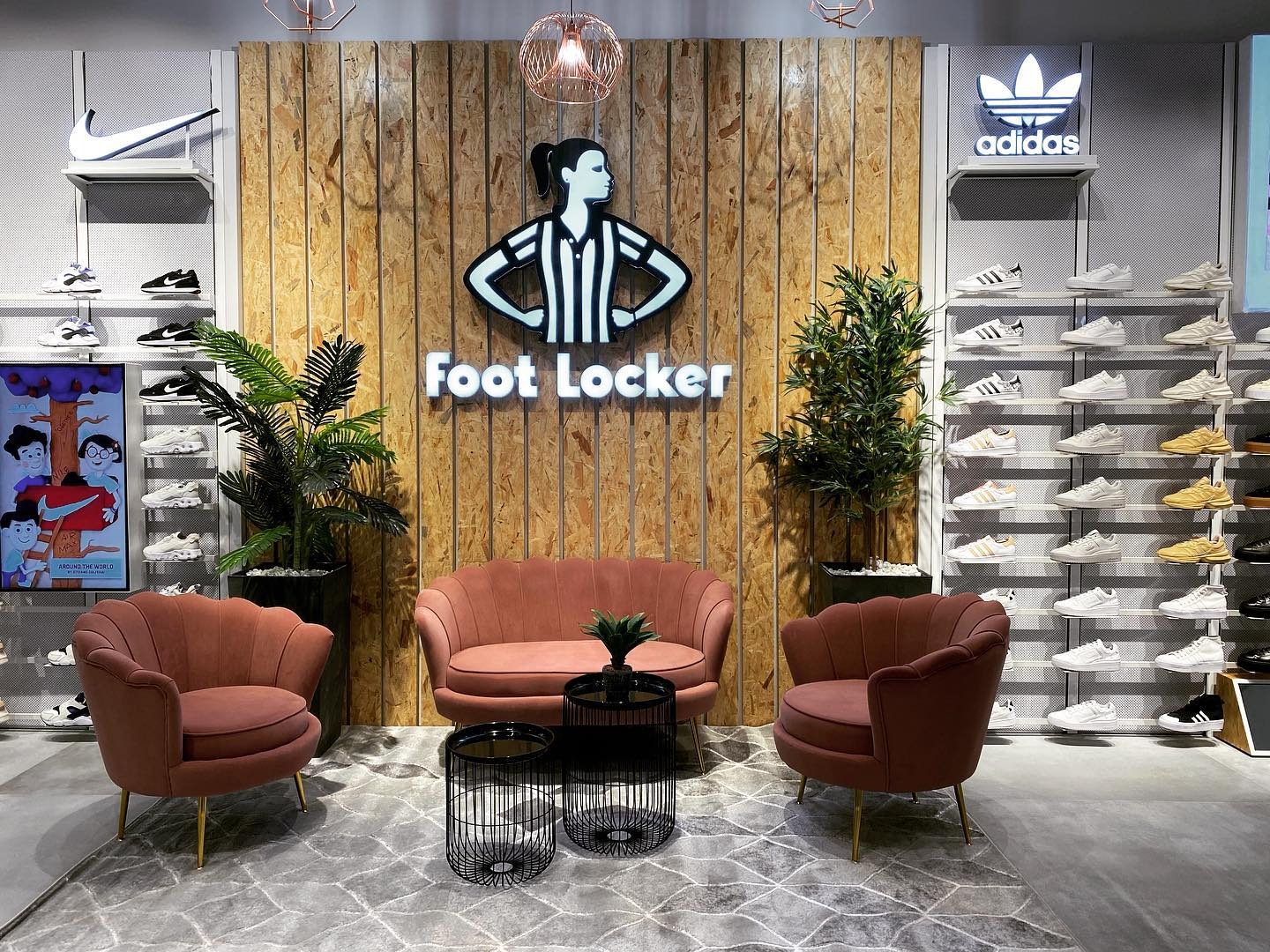 Wnętrze sklepu sportowego Foot Locker z logo podświetlanym na drewnianej ścianie, obok regały z butami Nike i Adidas, oraz wygodne fotele w kolorze brudnego różu.