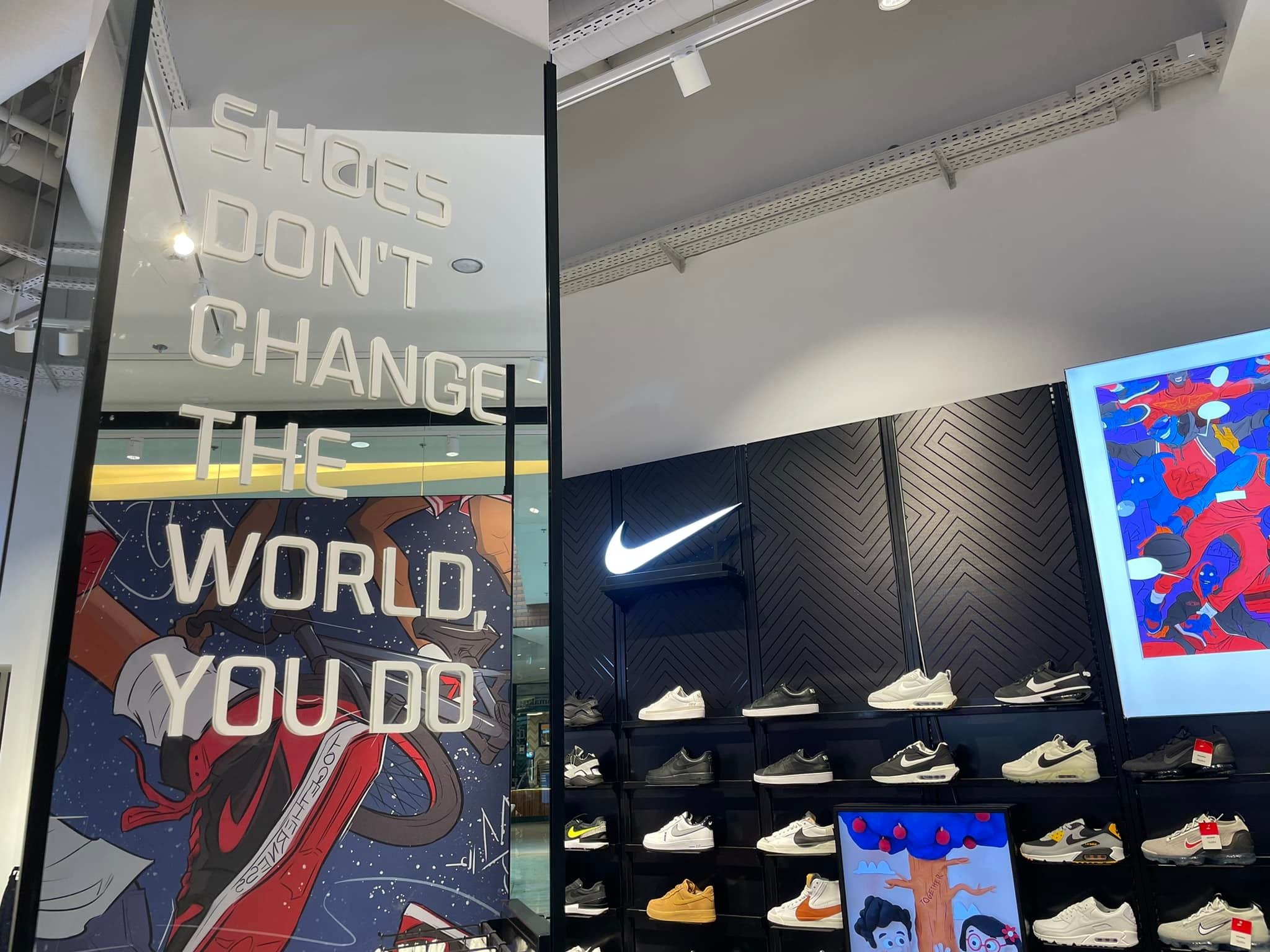 Wnętrze sklepu sportowego Nike z ekspozycją obuwia na półkach i dużym napisem reklamowym 'SHOES DON'T CHANGE THE WORLD, YOU DO' na szklanej witrynie.