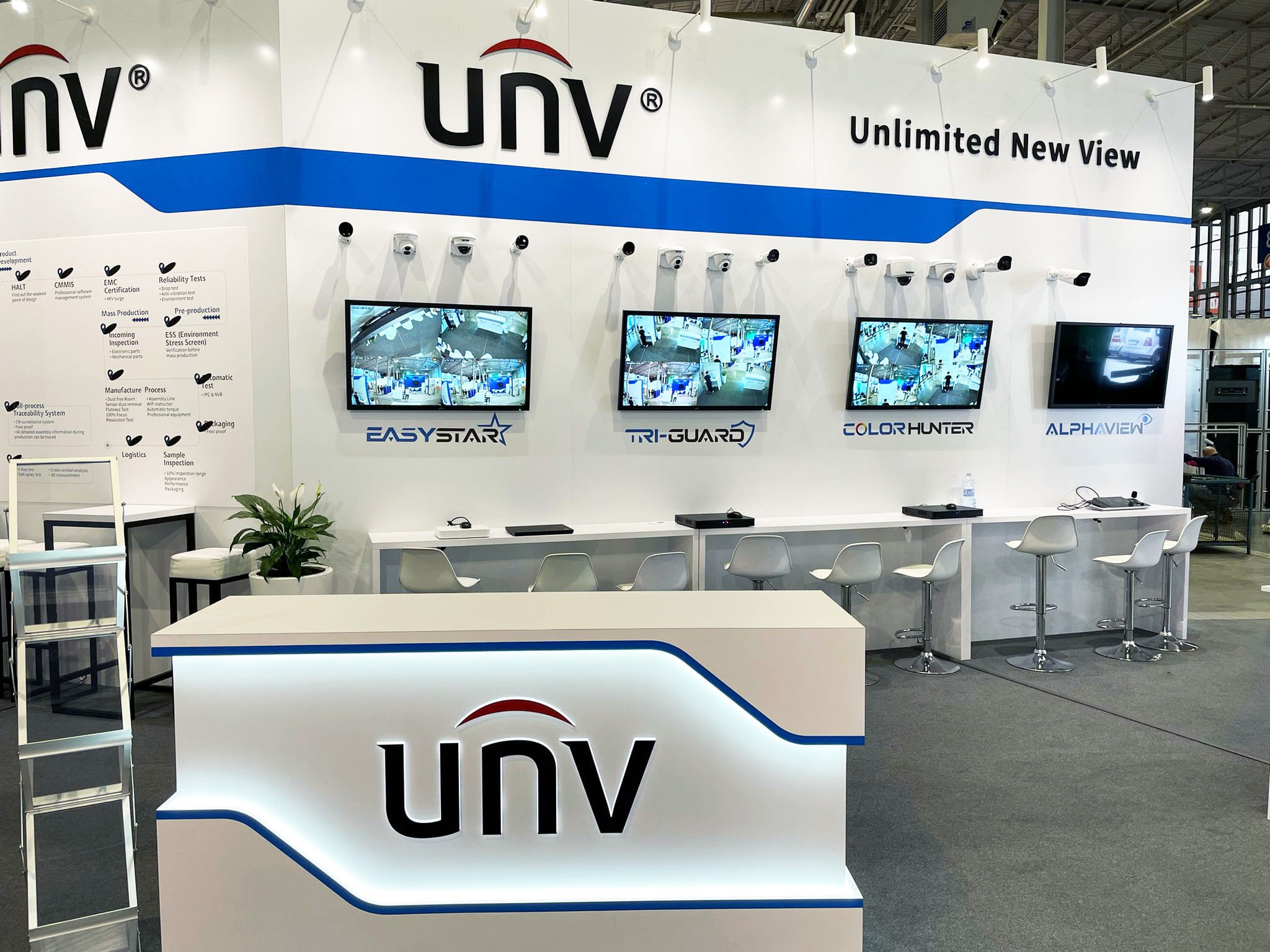 Stoisko targowe z logo firmy UNV, prezentujące systemy monitoringu wizyjnego z kamerami i monitorami wyświetlającymi obraz z kamer. Na ścianie tablica z opisem procesu produkcji i testów.