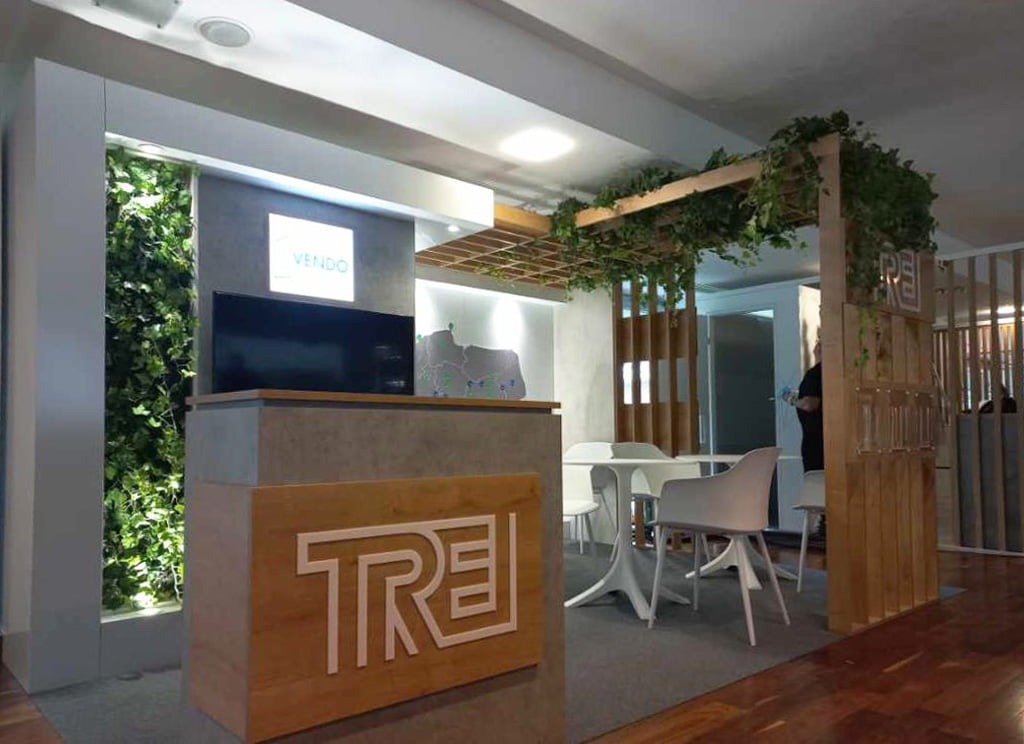 Aranżacja przestrzeni biurowej z recepcją z logo TRE, ścianą z żywej zieleni, drewnianą konstrukcją z roślinami pnącymi i stolikami z krzesłami w tle.