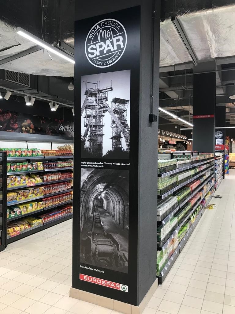 Wielkoformatowy wydruk na filarze w supermarkecie Eurospar przedstawiający czarno-białe zdjęcia szybów górniczych i wnętrza kopalni z podpisami 'Moja Okolica, Mój Spar, dumni z okolicy' oraz 'Szyby...